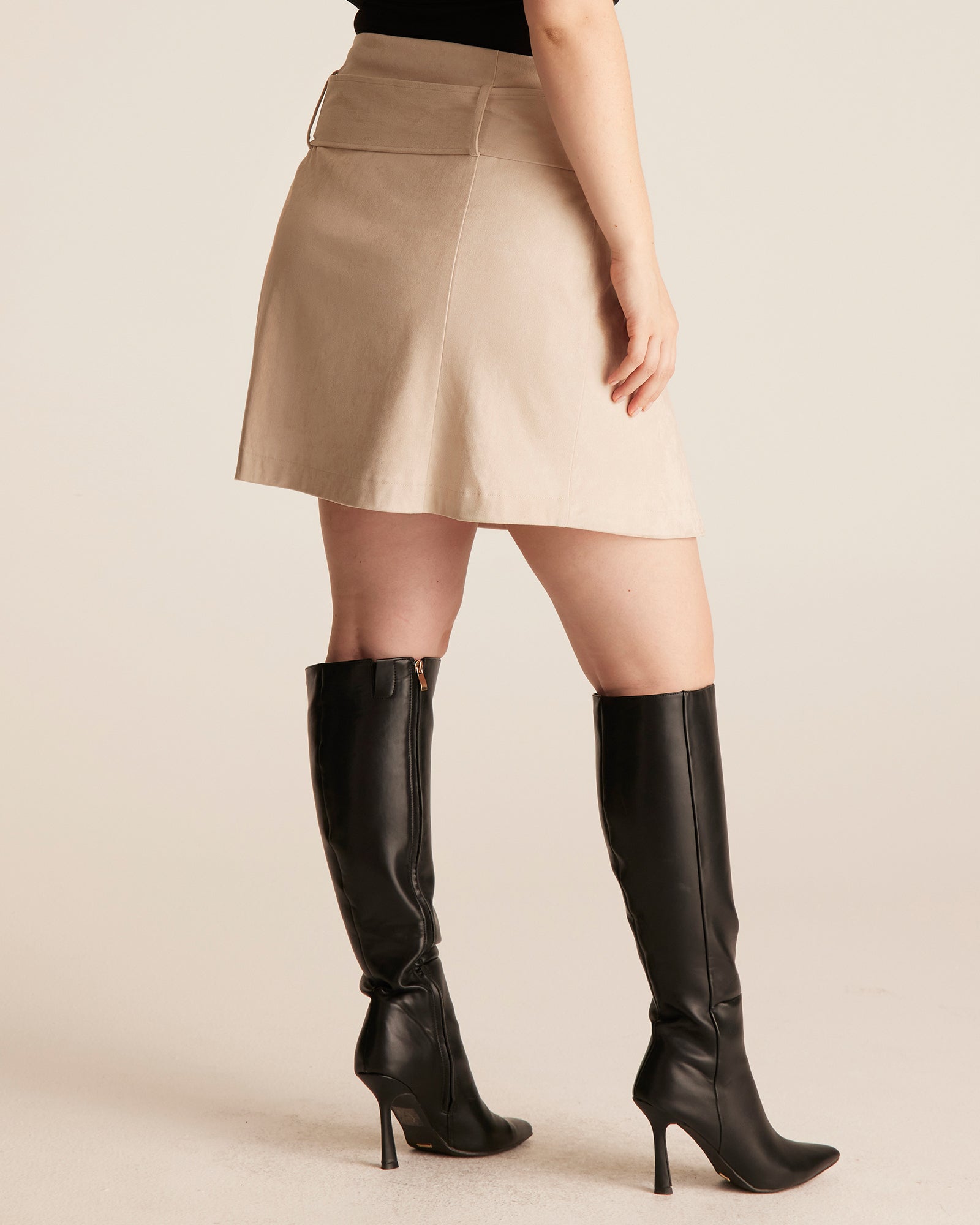 Person wearing a beige skirt and black knee-high boots on a beige background