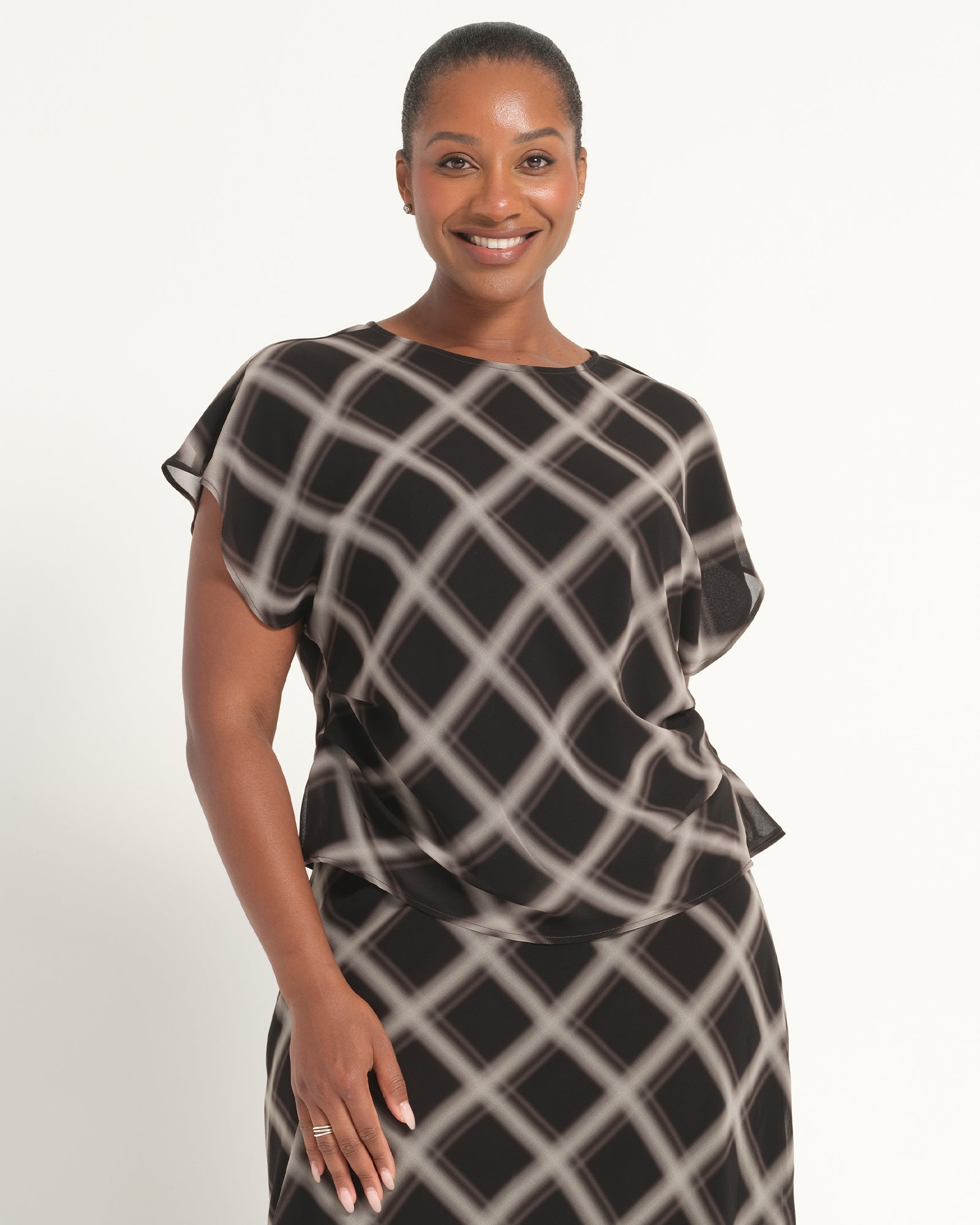 A woman modeling a black and beige geometric patterned dress with draped cap sleeves against a plain white studio background.