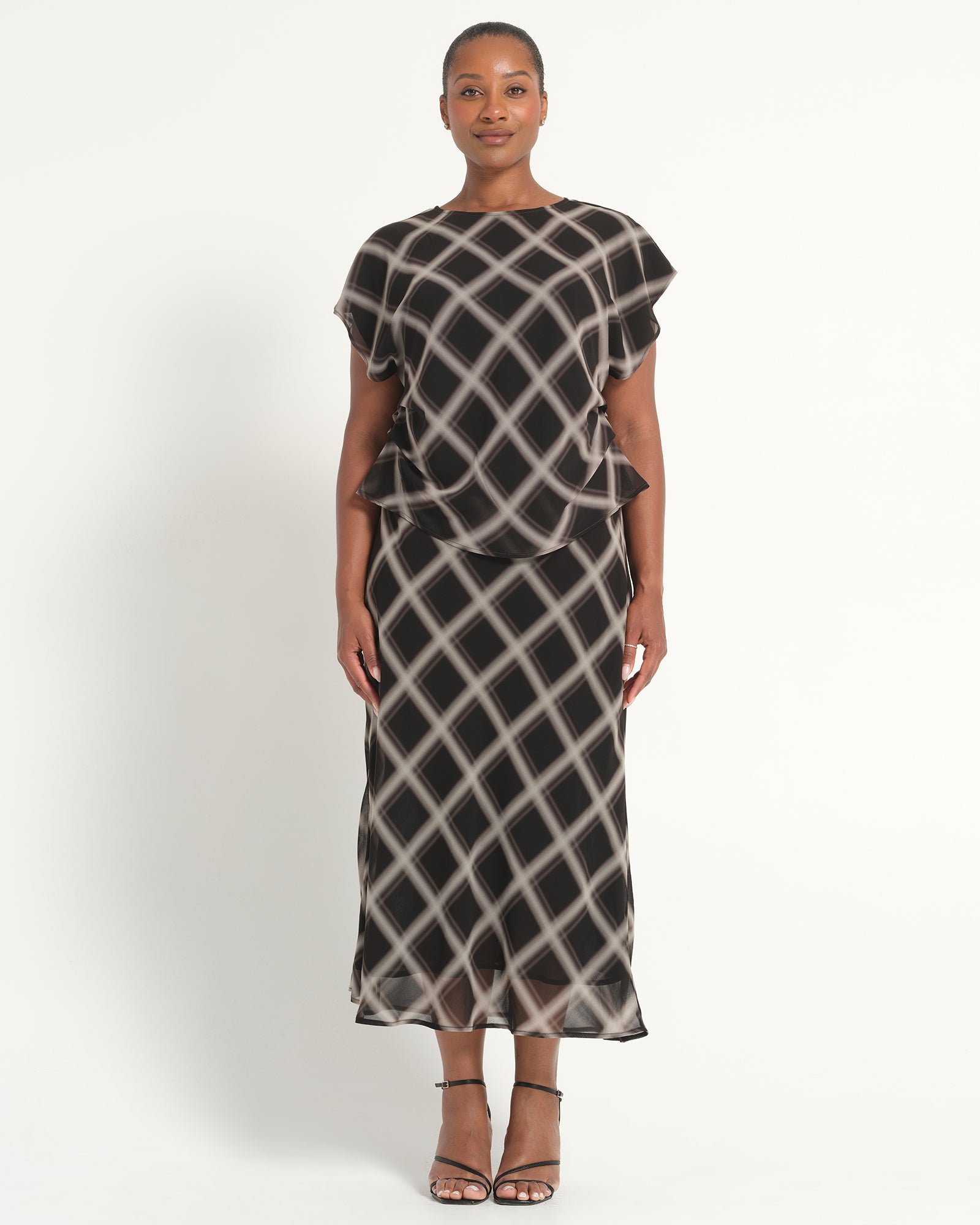 A woman modeling a sheer black midi dress with a beige geometric lattice pattern, featuring short flutter sleeves and a ruffled waist detail, set against a plain light background.