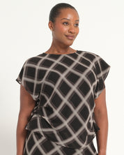 A woman wearing a black and white grid-patterned top with flutter sleeves, photographed against a plain white studio background.