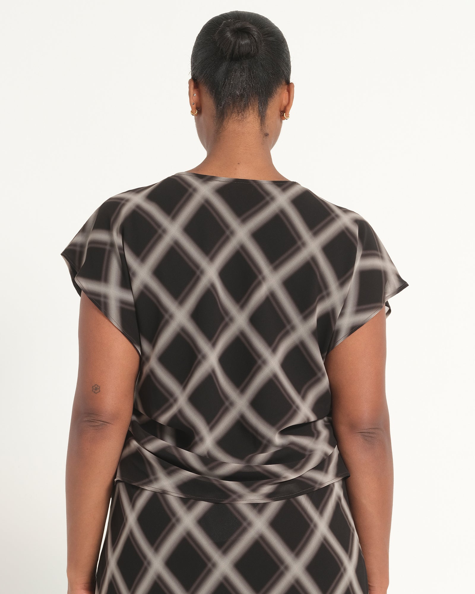 A woman viewed from the back wearing a short-sleeve black dress with a white diagonal lattice pattern, set against a plain white studio background.