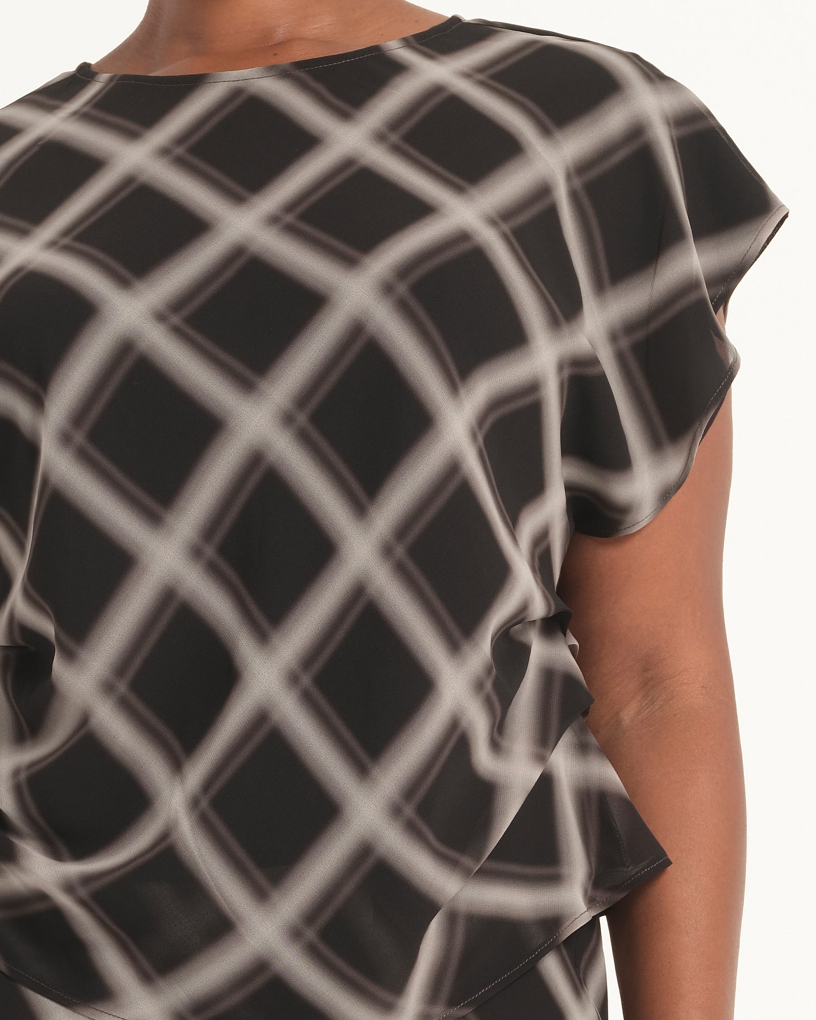 A close-up of a black short-sleeve blouse with a white grid pattern, worn by a model. The blouse features a relaxed fit, draped sleeves, and an asymmetrical ruffled hem.