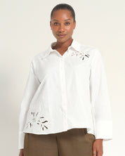 A woman modeling a white long-sleeve button-up shirt with eyelet embroidery on the chest and lower front, paired with brown pants, against a plain light background.