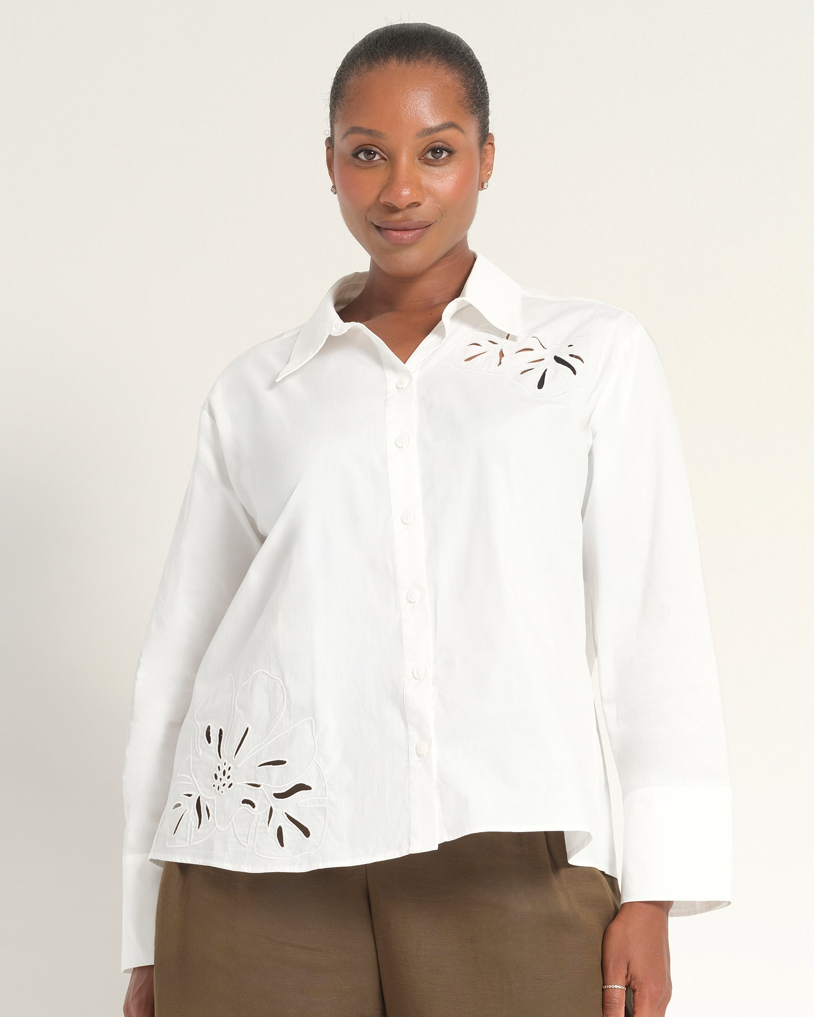 A woman modeling a white long-sleeve button-up shirt with eyelet embroidery on the chest and lower front, paired with brown pants, against a plain light background.