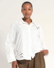 A woman modeling a white long-sleeve button-up shirt with decorative cutout embroidery on the chest and lower hem, paired with brown trousers, against a plain light beige background.