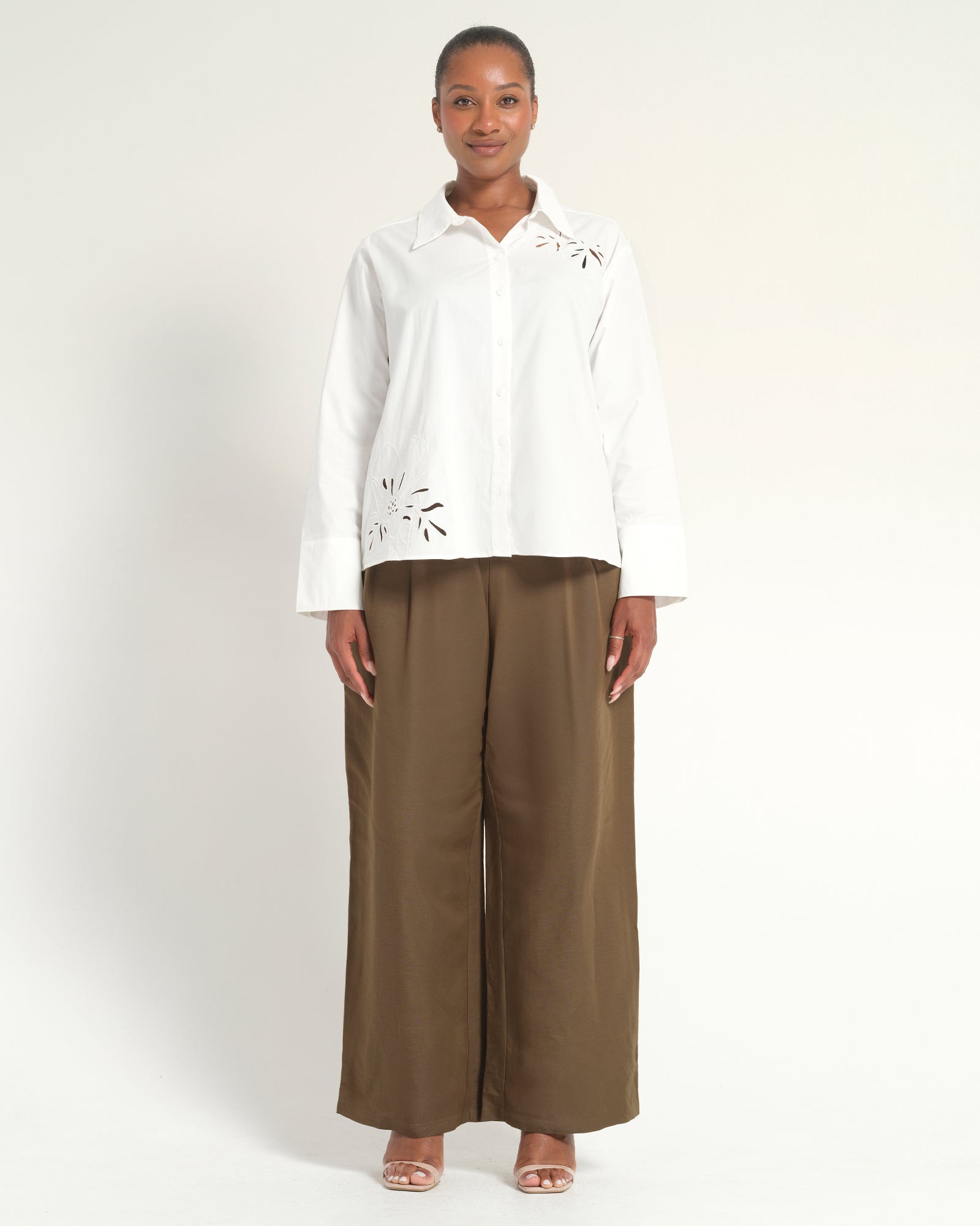 A woman stands against a plain light background wearing a white long-sleeve button-up blouse with eyelet cutout details on the shoulders and lower front, paired with wide-leg brown pants and nude heeled sandals.