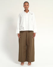 A woman stands against a plain light background wearing a white long-sleeve button-up blouse with eyelet cutout details on the shoulders and lower front, paired with wide-leg brown pants and nude heeled sandals.