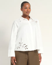 A woman modeling a white long-sleeve button-up blouse with decorative cutout embroidery on the chest and lower front, paired with brown pants, against a plain light background.