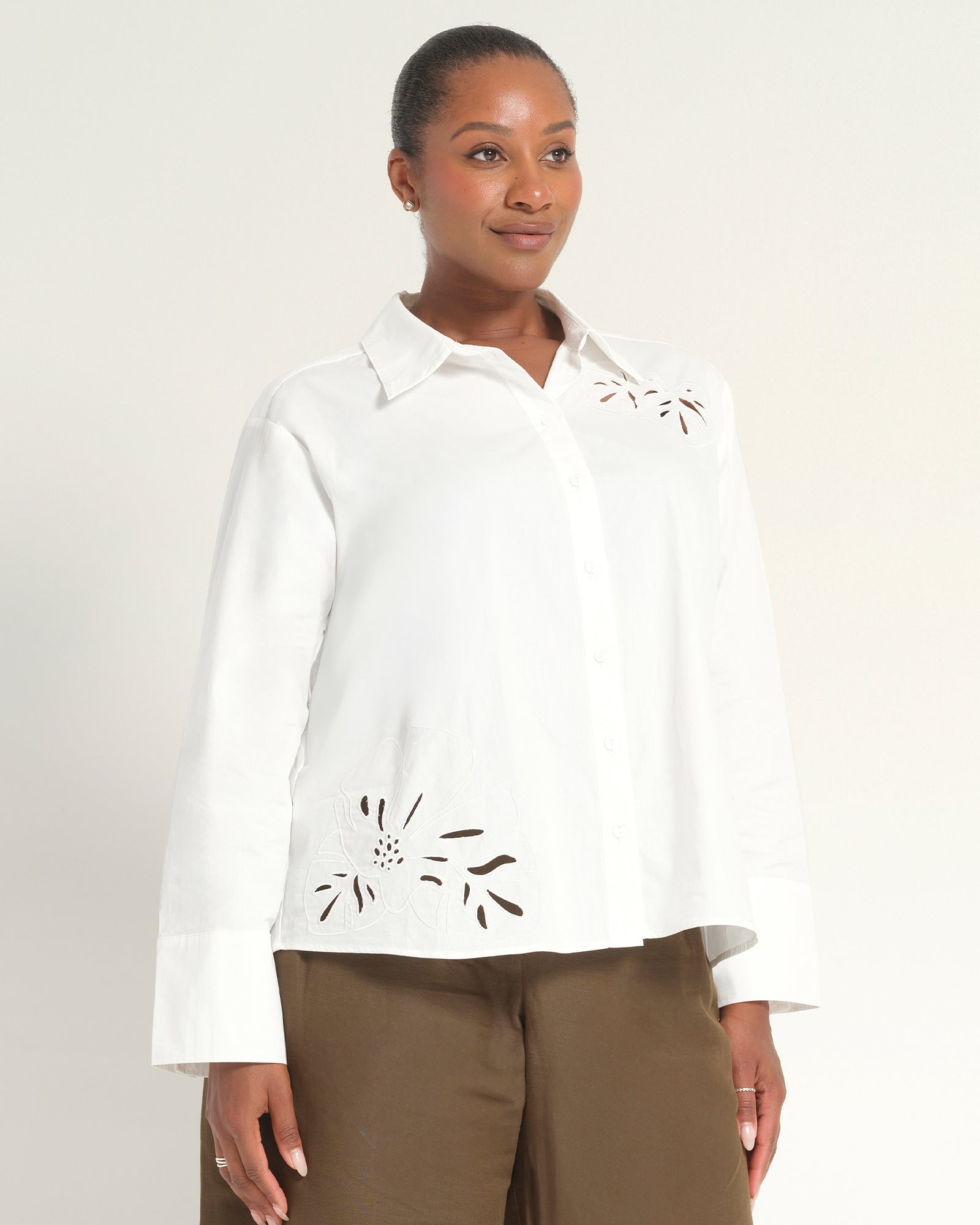 A woman modeling a white long-sleeve button-up blouse with decorative cutout embroidery on the chest and lower front, paired with brown pants, against a plain light background.
