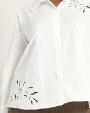 A close-up of a white button-front shirt with floral eyelet embroidery on the lower left side and upper chest, worn with brown bottoms against a light neutral background.