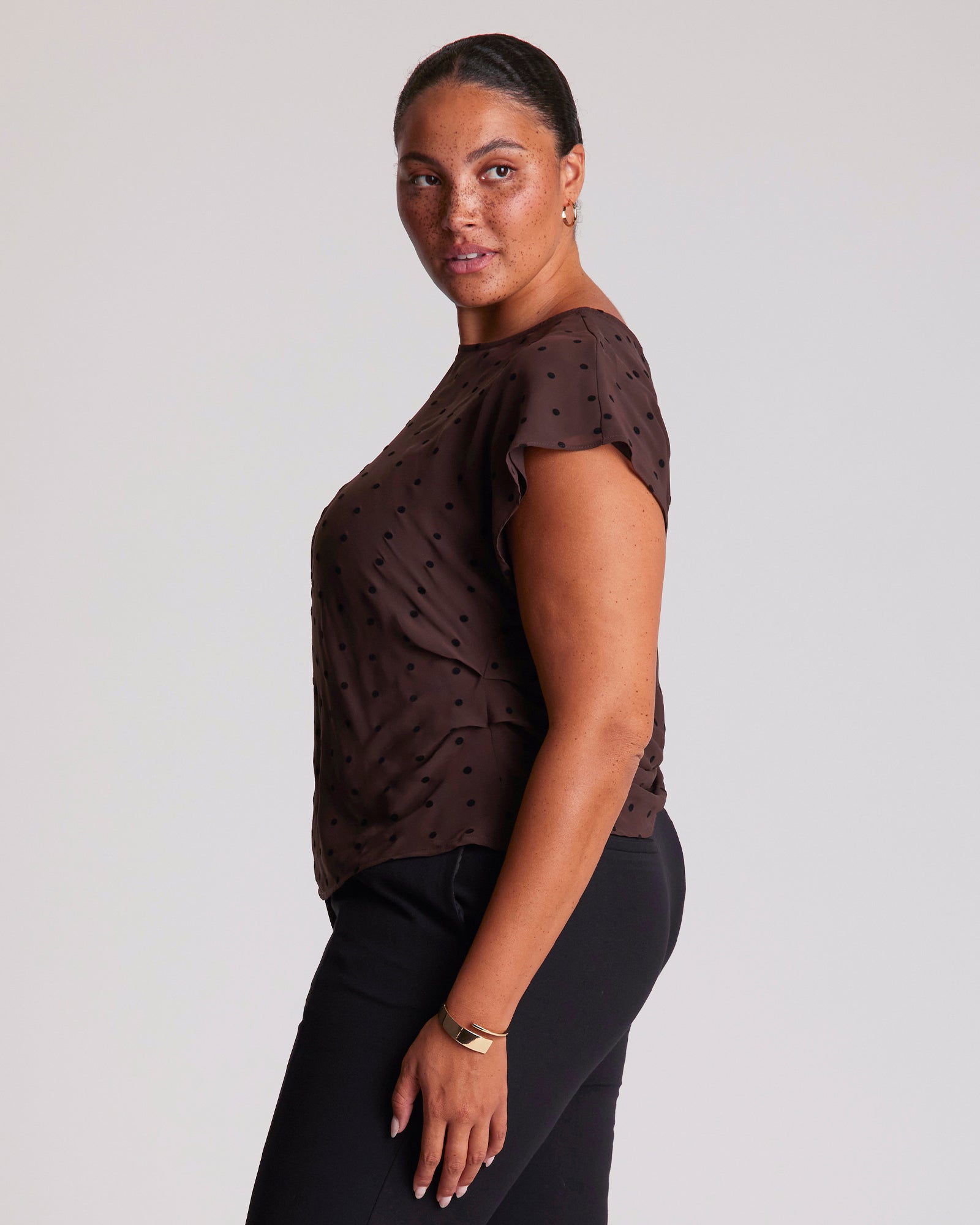 Woman wearing a brown top with black polka dots and black pants on a light gray background