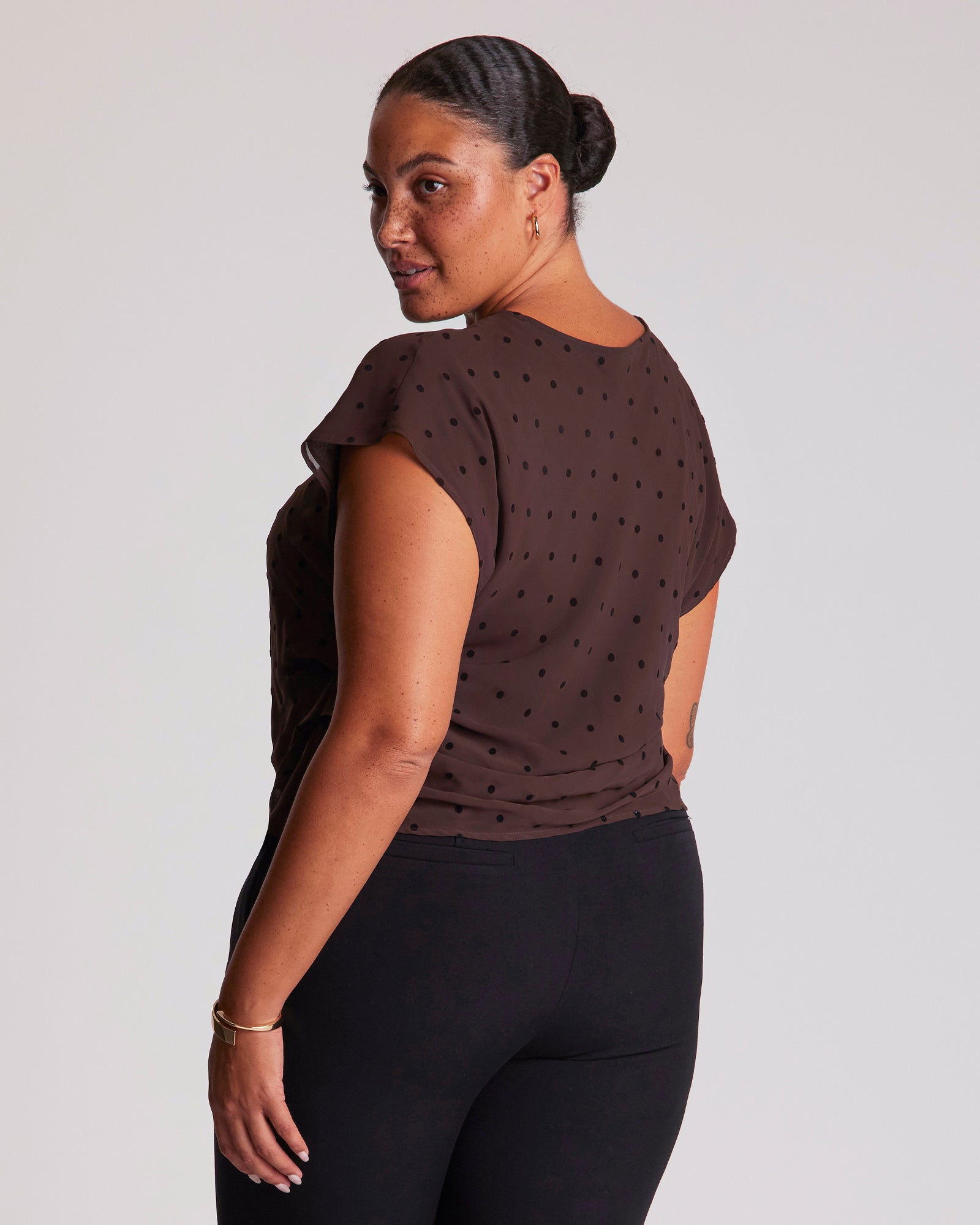 Woman wearing a brown polka dot top and black pants on a light gray background