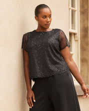 Woman wearing a black sequin top and black pants against a beige wall.