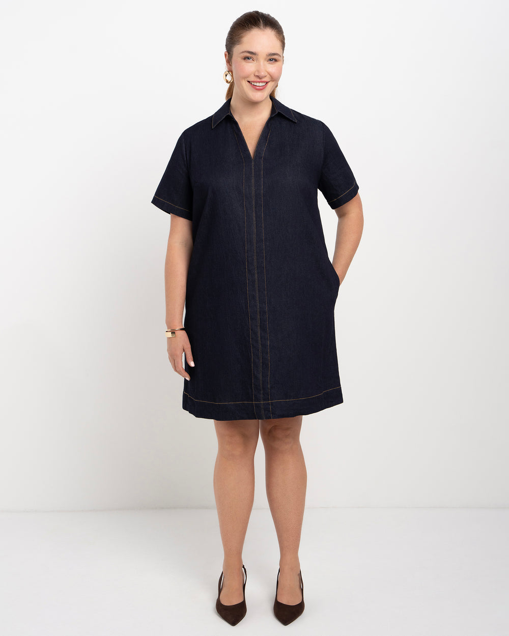 A woman modeling a dark navy blue short-sleeve collared shirt dress with contrast stitching in tan, standing against a plain white studio background.