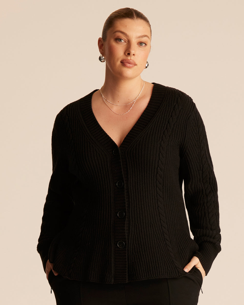 Woman wearing a black ribbed cable knit cardigan with a button up front and V-neck design against a beige background