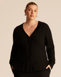 Woman wearing a black ribbed cable knit cardigan with a button up front and V-neck design against a beige background