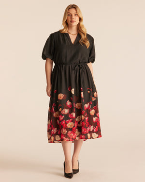 A woman modeling a black midi dress with short puff sleeves, a V-neckline, and a red and beige tulip print concentrated along the lower skirt of a black dress, photographed against a plain beige studio background.