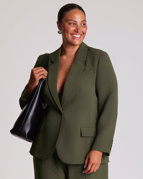 Woman wearing a dark green suit blazer, holding a black bag against a plain background.