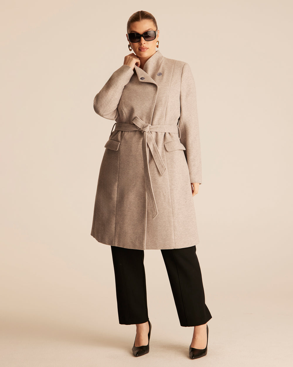Woman wearing a beige trench style coat with an asymmetrical funnel style neckline on a beige background with black tailored pants.