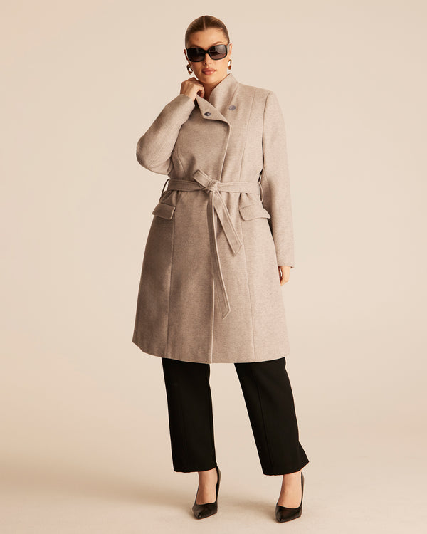 Woman wearing a beige trench style coat with an asymmetrical funnel style neckline on a beige background with black tailored pants.