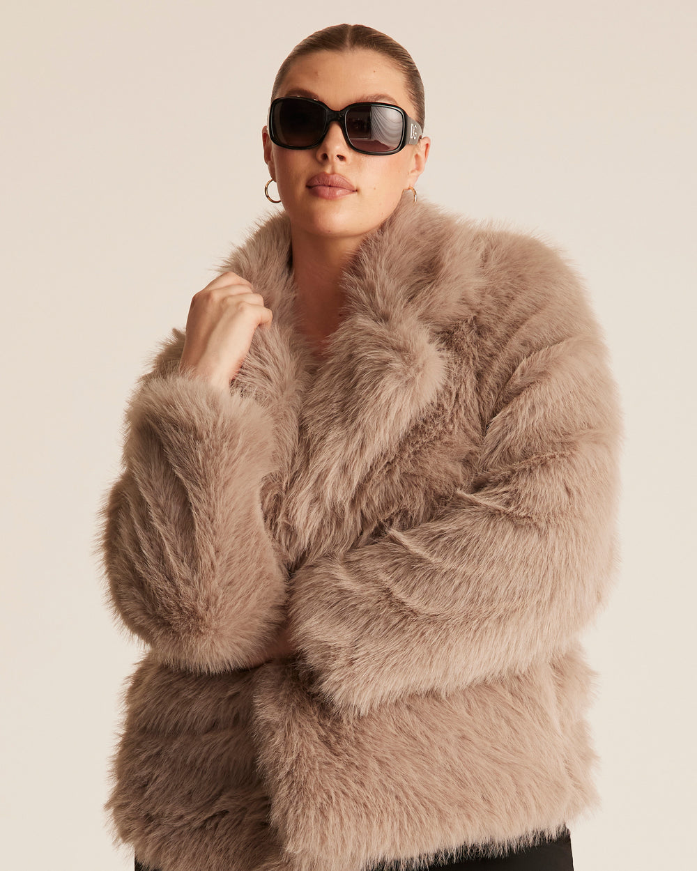 Woman wearing a fluffy faux fur jacket style coat in a pebble brown beige colour. Styled with sunglasses and a gold hoop earring on a plain background.