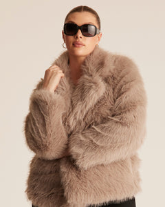 Woman wearing a fluffy faux fur jacket style coat in a pebble brown beige colour. Styled with sunglasses and a gold hoop earring on a plain background.