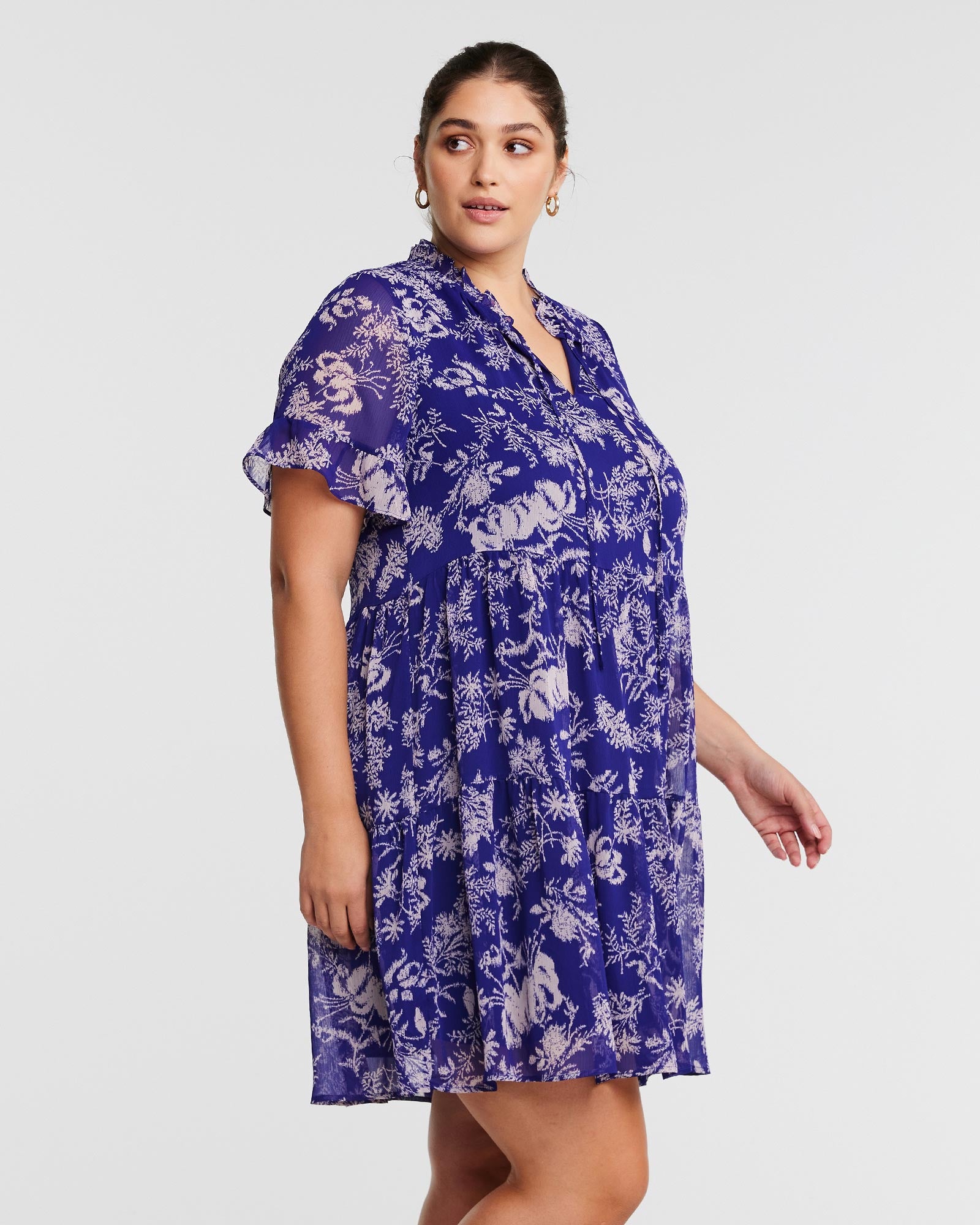 A plus size woman wearing an Estelle Ultra Leaf Purple Tiered Knee Length Dress with a blue floral print.