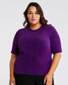 A woman wearing an Estelle Nora Fuzzy Purple Short-Sleeved Sweater and black pants.