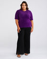 A plus size woman wearing an Estelle Nora Fuzzy Purple Short-Sleeved Sweater and black wide leg pants.