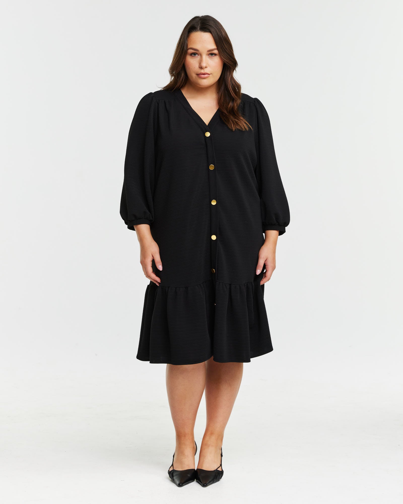 A plus size woman wearing the Elissa Dress, a black button down dress with gold hardware detailing.