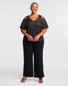 A woman wearing an Estelle Gold Dust Cold Shoulder Top and black wide leg pants.